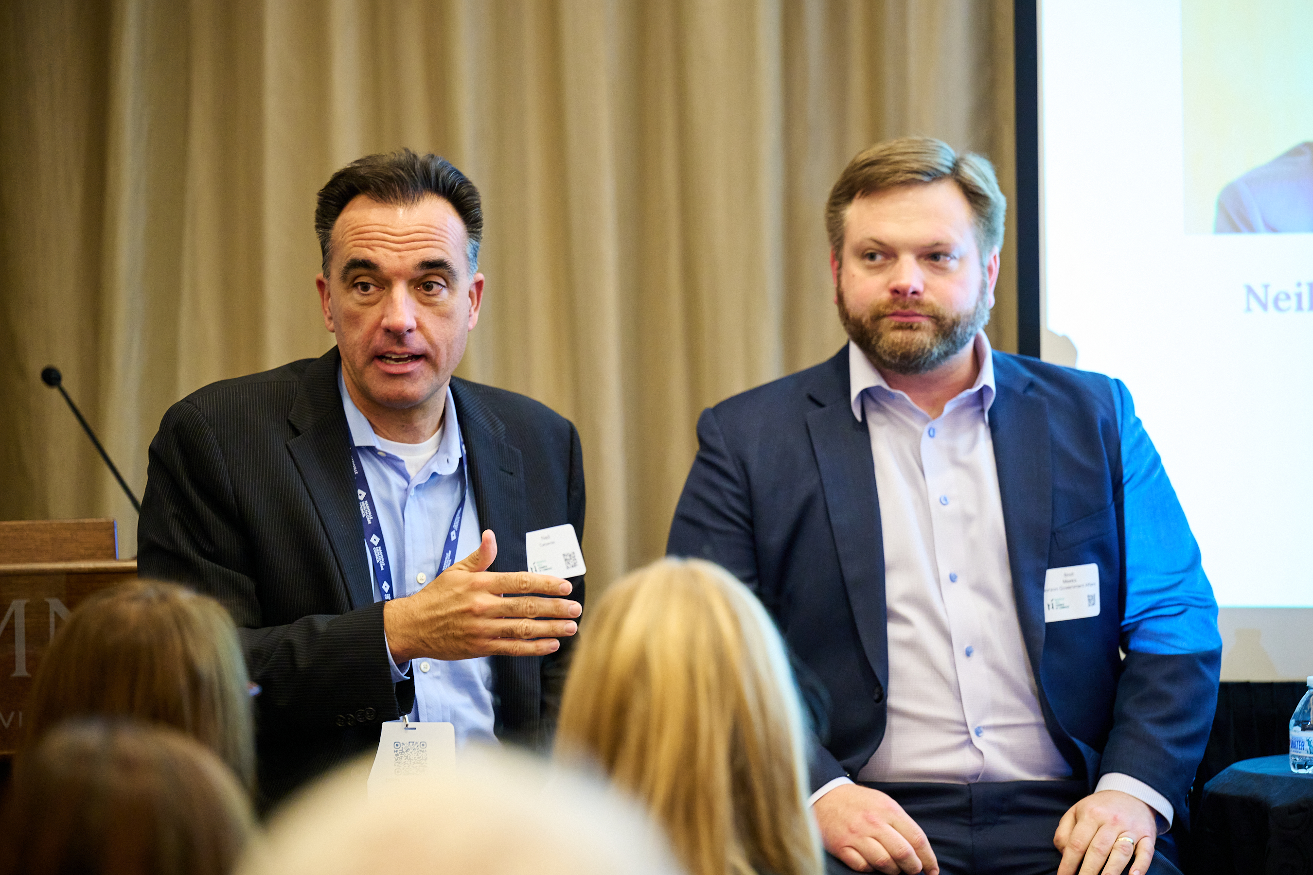 Conference panel discussion photo showing two professional men at a conference - one in a dark striped suit speaking on the left, and one in a navy suit on the right, both wearing name tags with an audience visible from behind in the foreground.